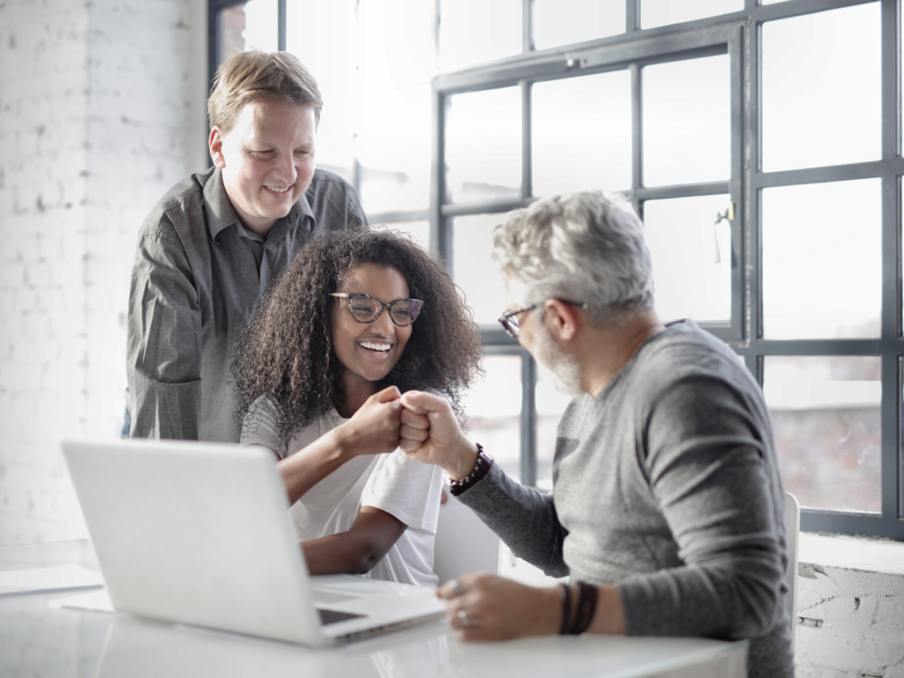 Drei Personen im Büro arbeiten gemeinsam an einem Laptop und geben sich motiviert einen Fistbump – Symbol für Teamzusammenhalt und positive Zusammenarbeit im Startup.